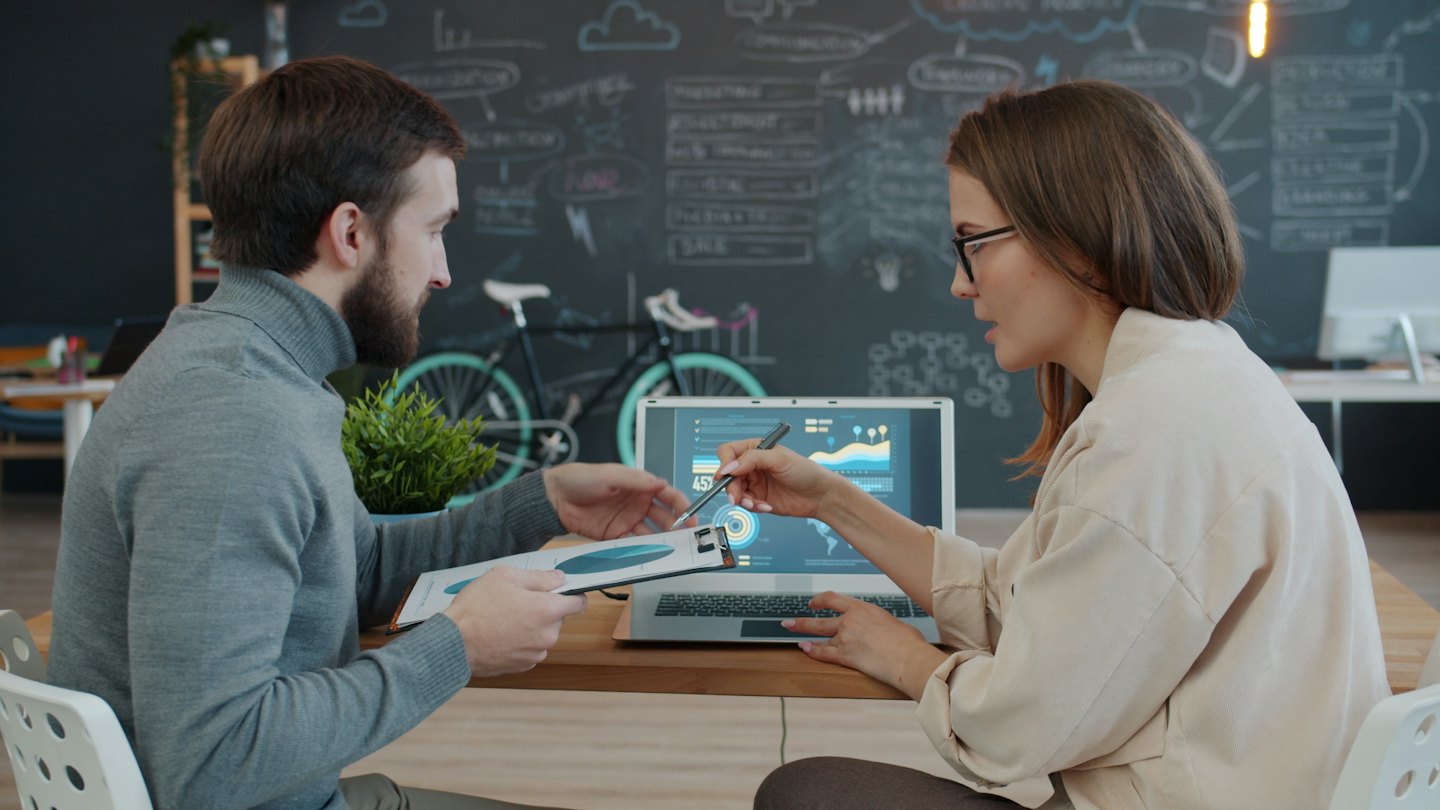 Dos personas de negocios discuten datos en una computadora portátil y un documento.
